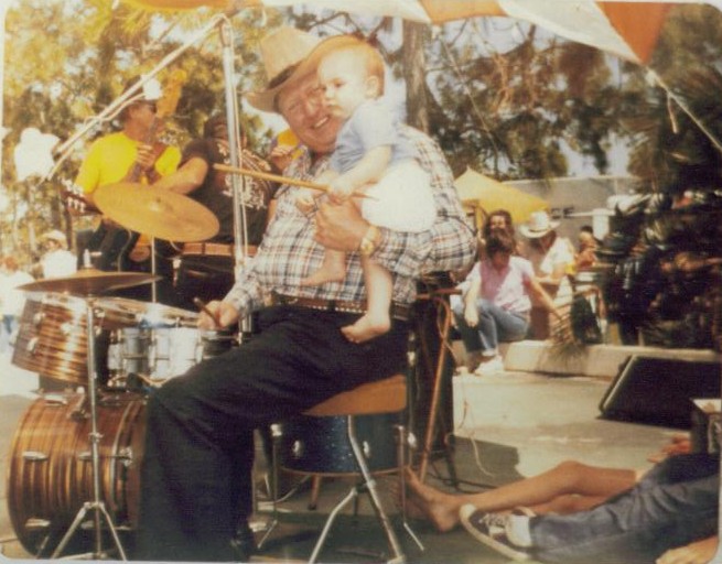 Baby Randy on a drummer's lap behind the kit at an outdoor show
