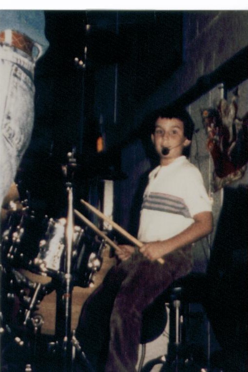 Randy as a kid playing drums on stage with a headset mic